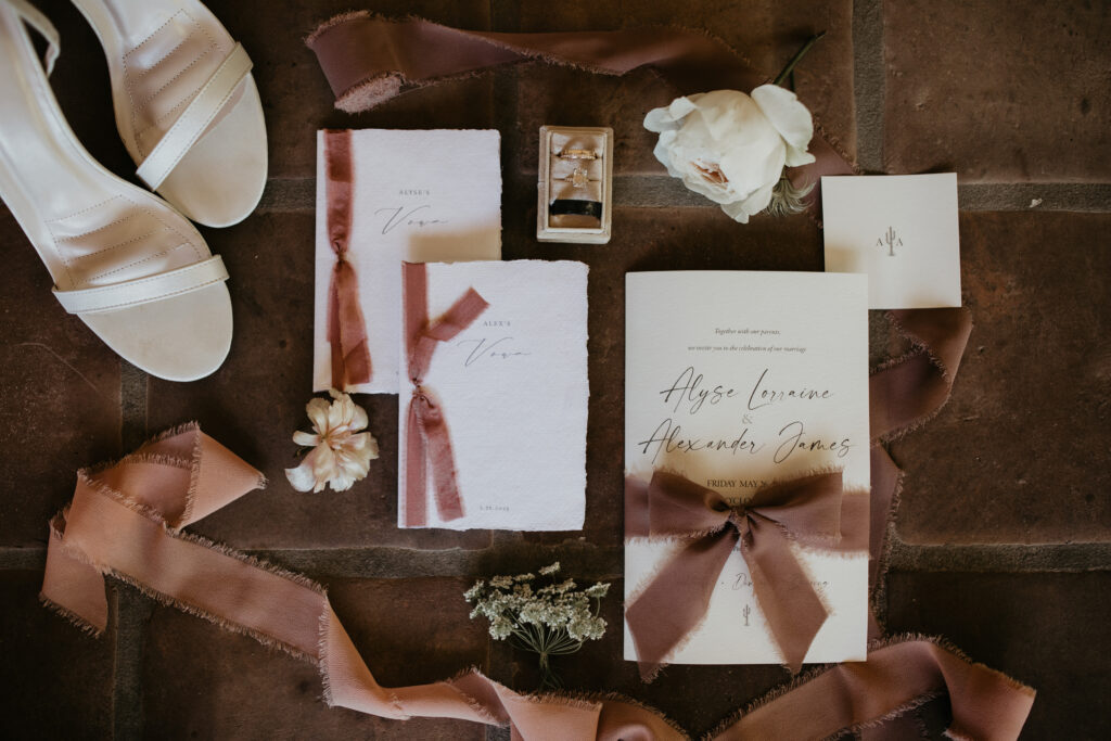 A flat lay photo that includes the brides shoes, rings, vow books, invitation. There are two different ribbons, one is a light pink, and the other is a mauve. There are florals laid around the image that are from the bride's bouquet.